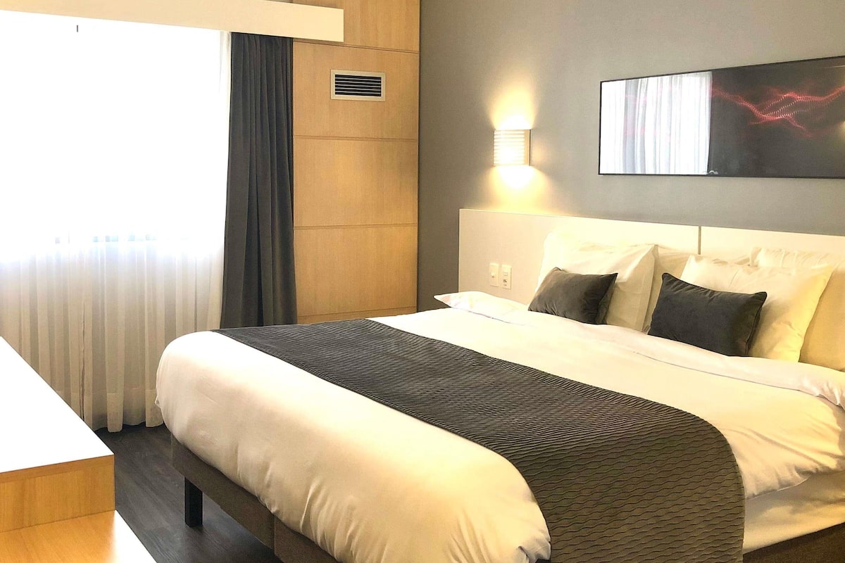 Ein Hotelzimmer mit einem großen Bett, einem Nachttisch, einer Wandleuchte und einem Fenster mit Vorhängen.