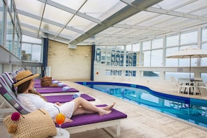 Un área junto a la piscina con sillones de descanso, una piscina cubierta con paredes de vidrio y vistas a la ciudad.