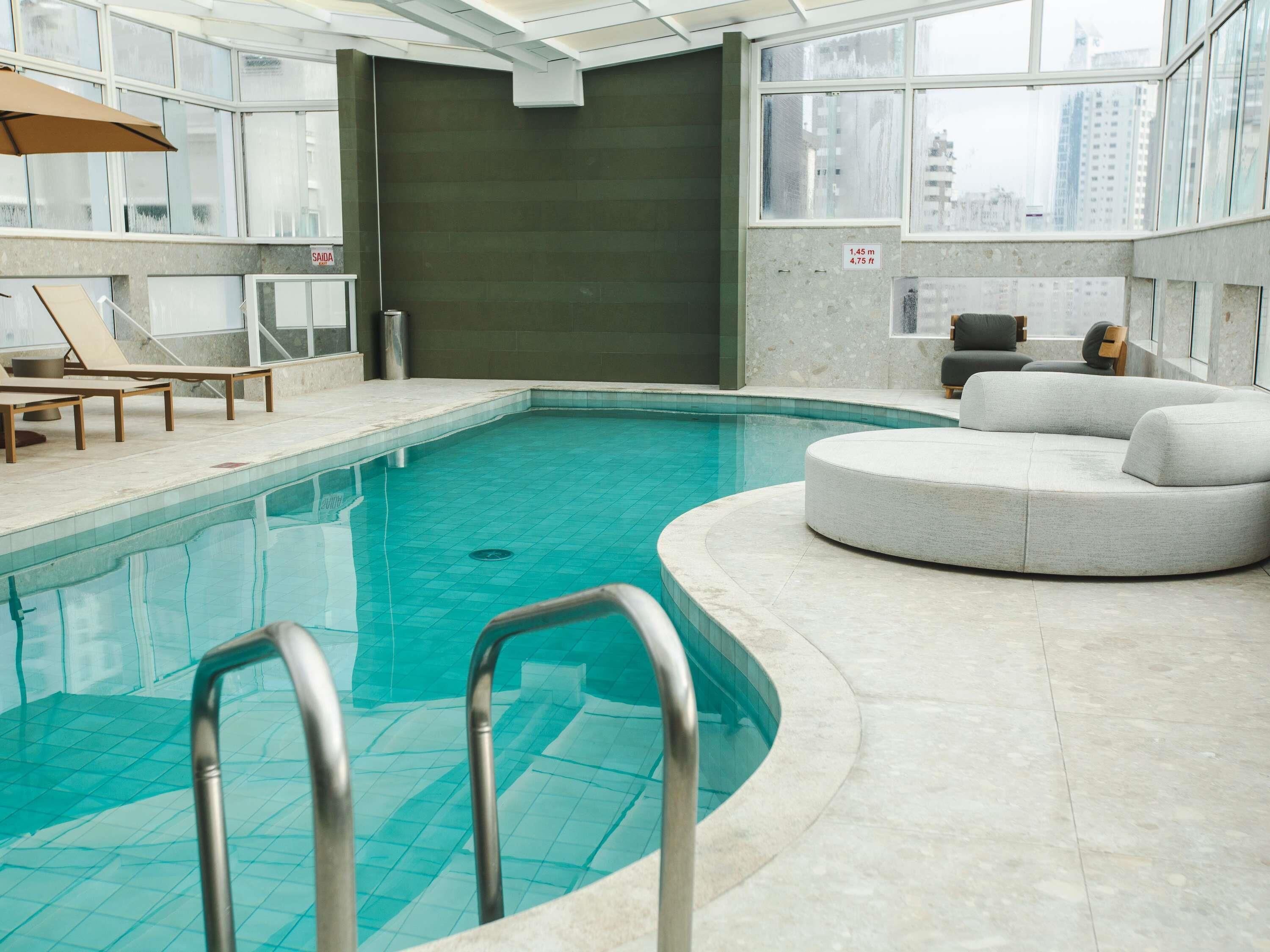 An indoor swimming pool with a curved edge, a lounge area with chairs, and a view of the city through large windows.