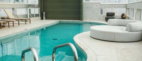 An indoor swimming pool with a curved edge, a lounge area with chairs, and a view of the city through large windows.