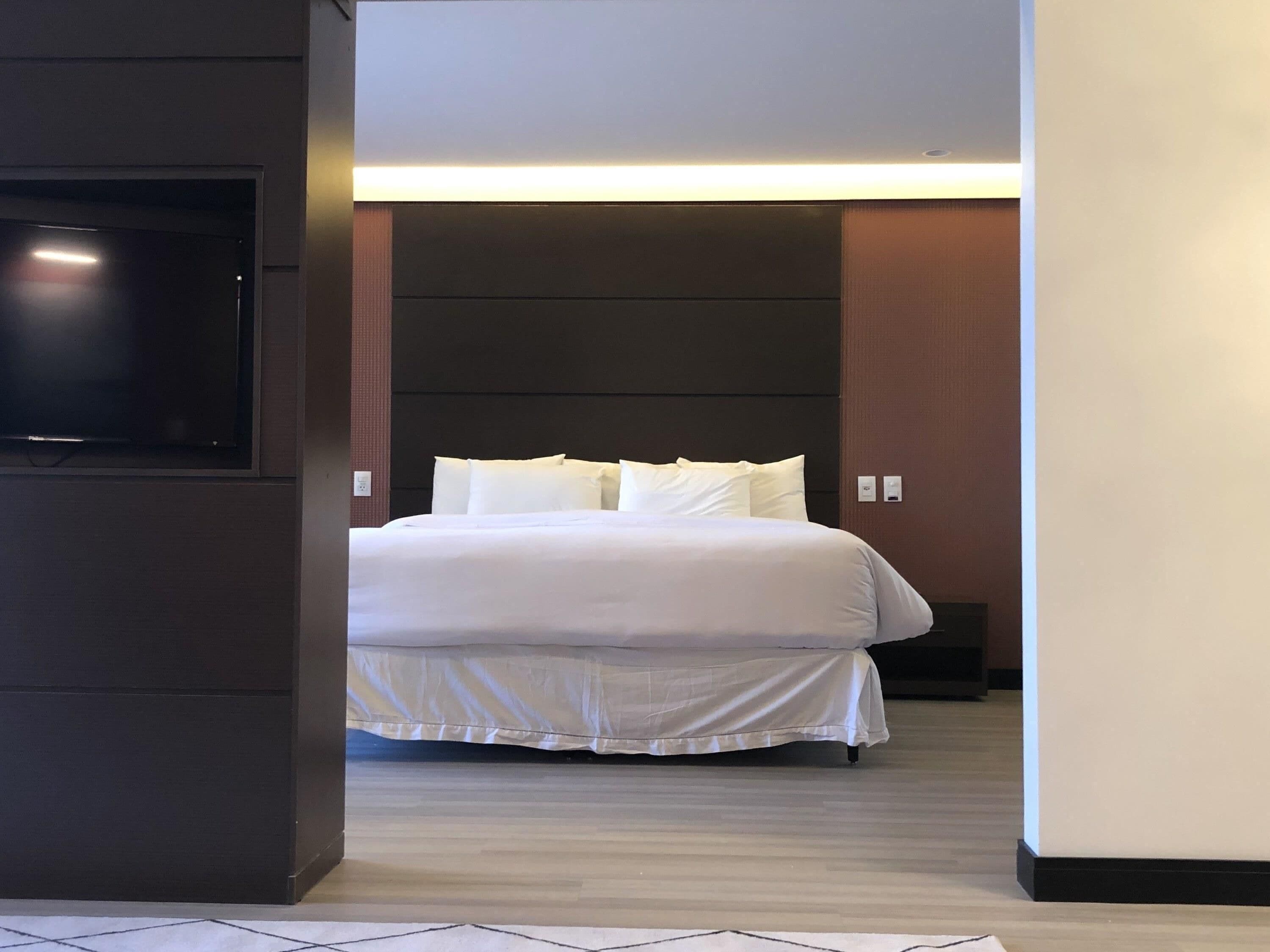 A modern hotel room with a large bed, a flat-screen TV mounted on the wall, and a bedside table.