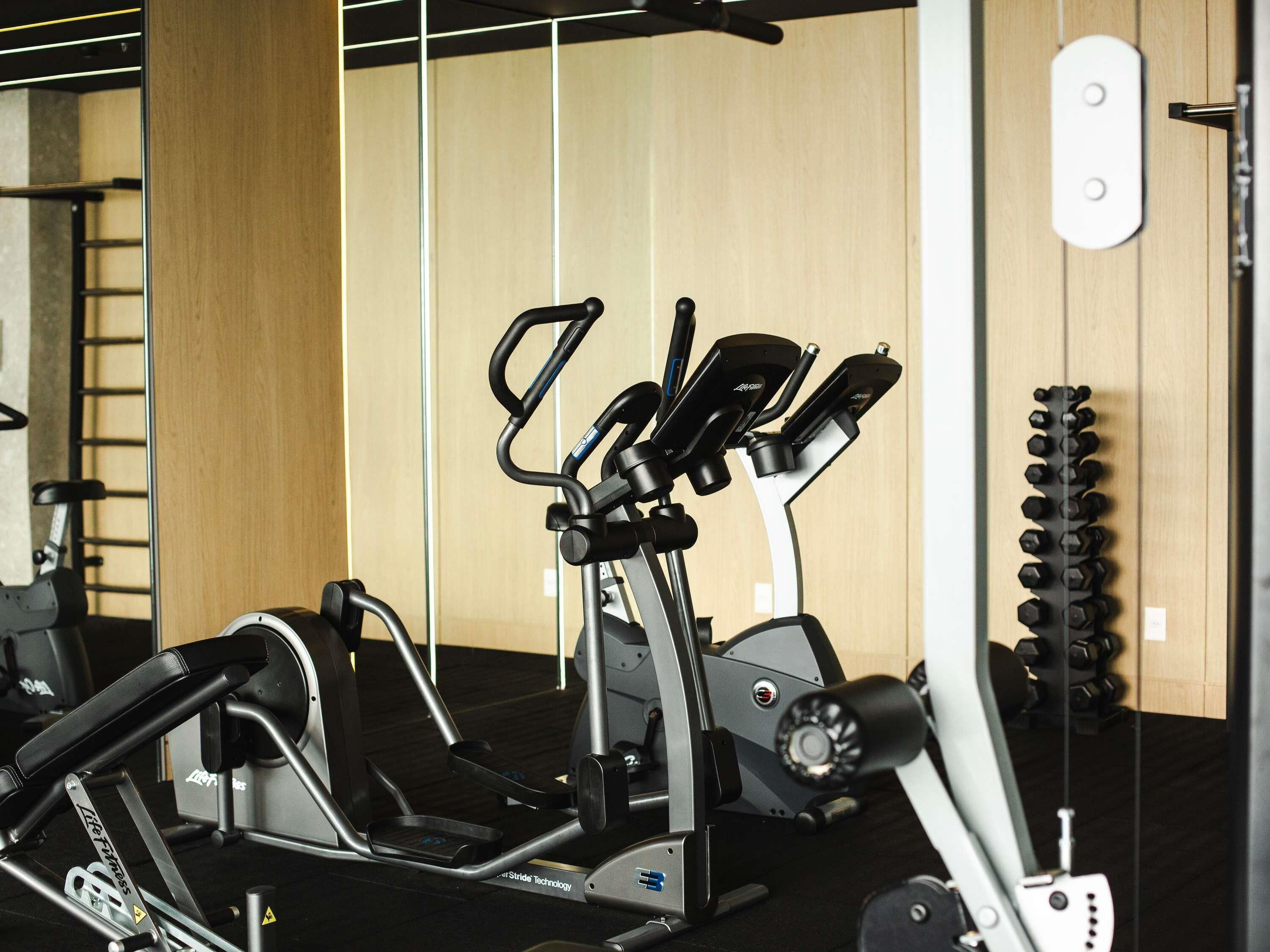 A stationary bike with a digital display, a wall-mounted rack with dumbbells, and a wooden panel wall.