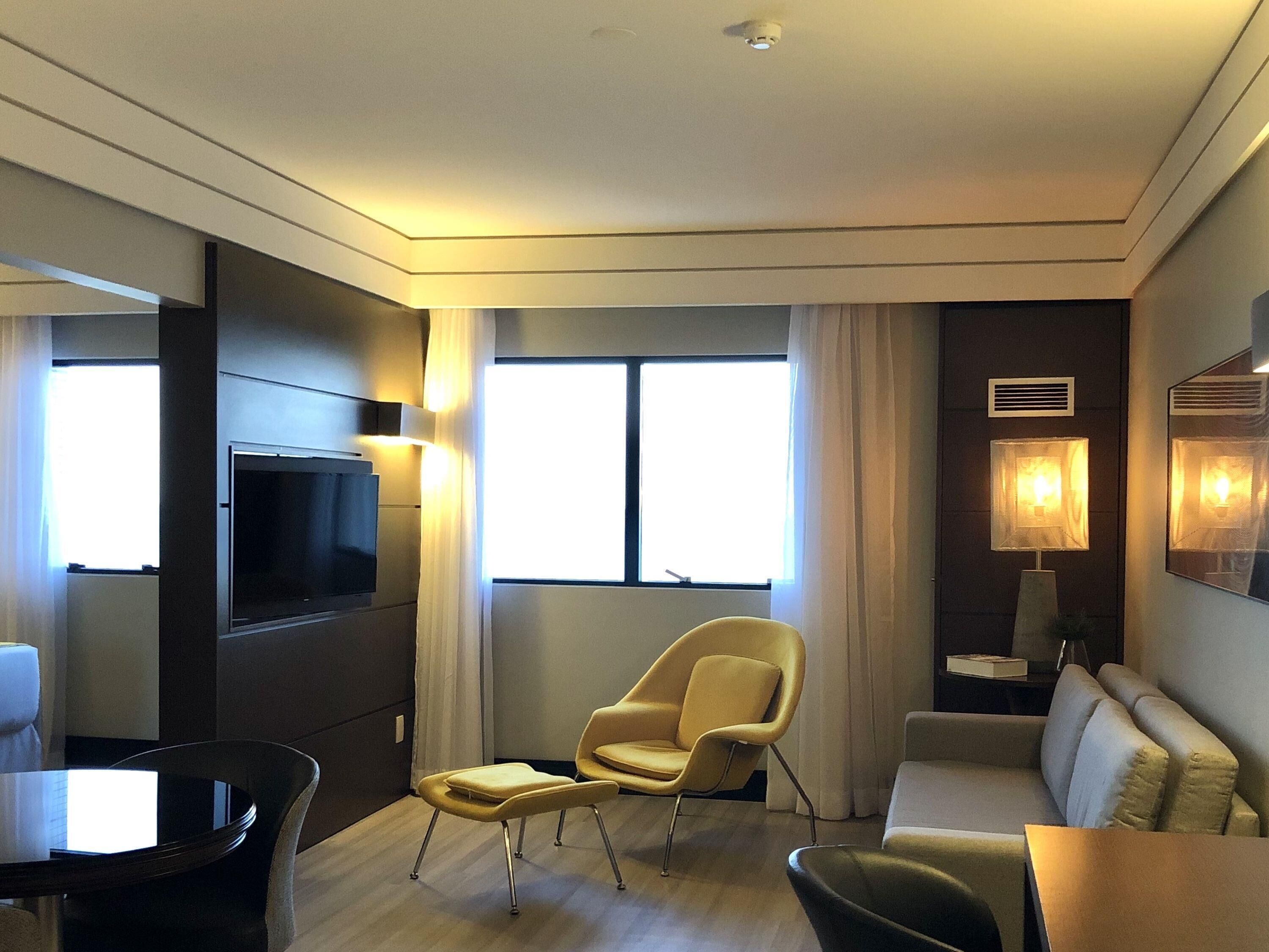 Quarto de hotel moderno com televisão de tela plana, sofá, poltrona, mesa pequena e abajur.