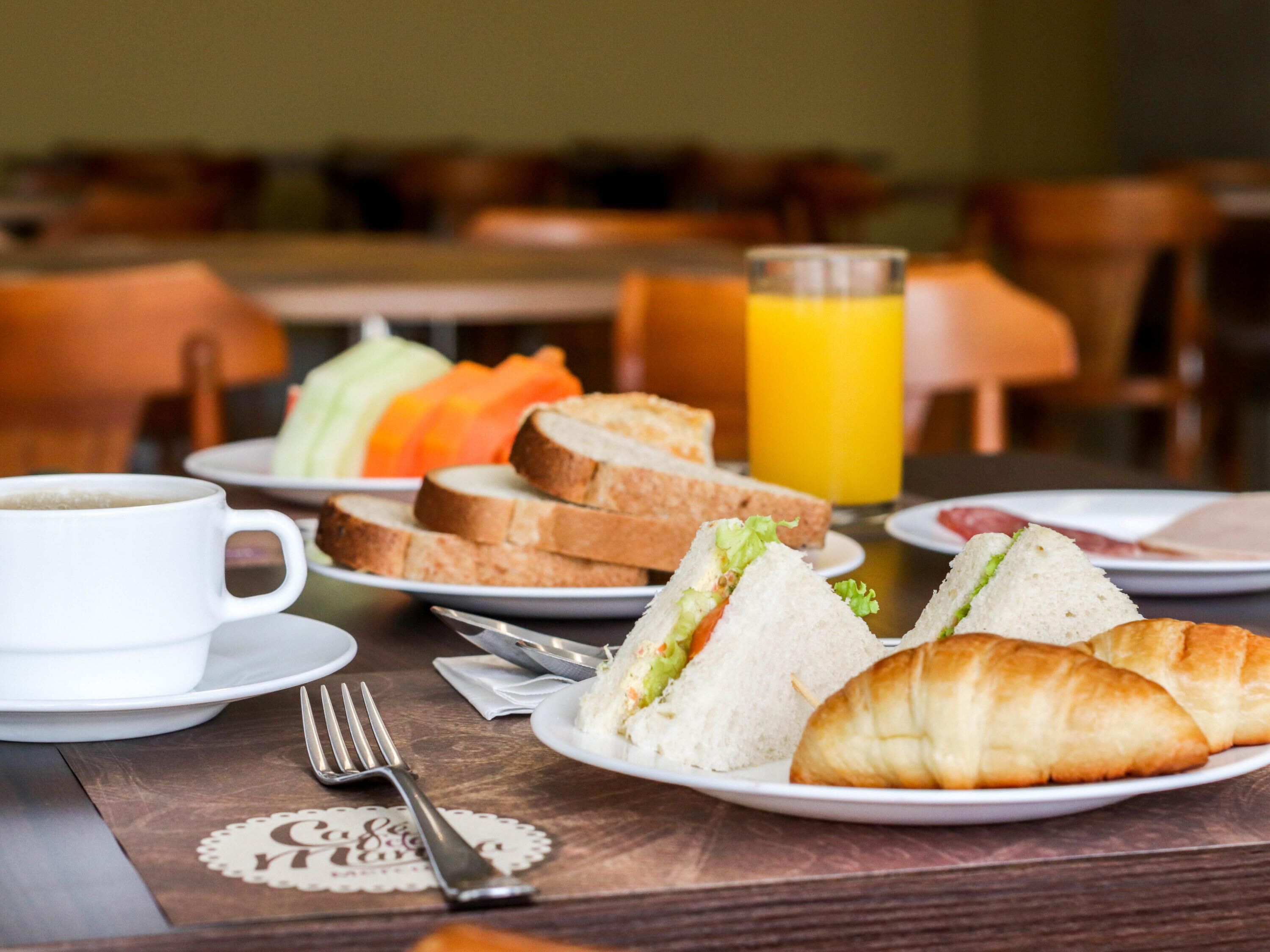 A breakfast setting with a cup, sandwiches, croissants, and a glass of orange juice on a table.