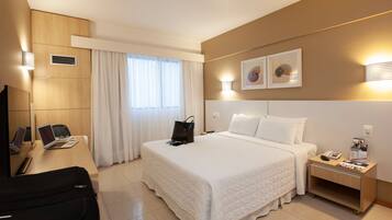 Quarto de hotel com cama, escrivaninha com um laptop, cadeira, mala, mesa de cabeceira com telefone e luminária fixada na parede.