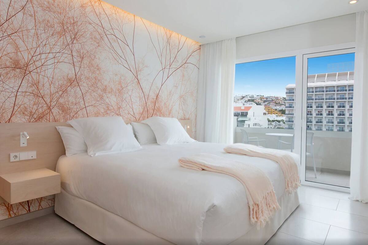 A modern bedroom with a large bed, a nightstand, and a view of the city through a balcony.