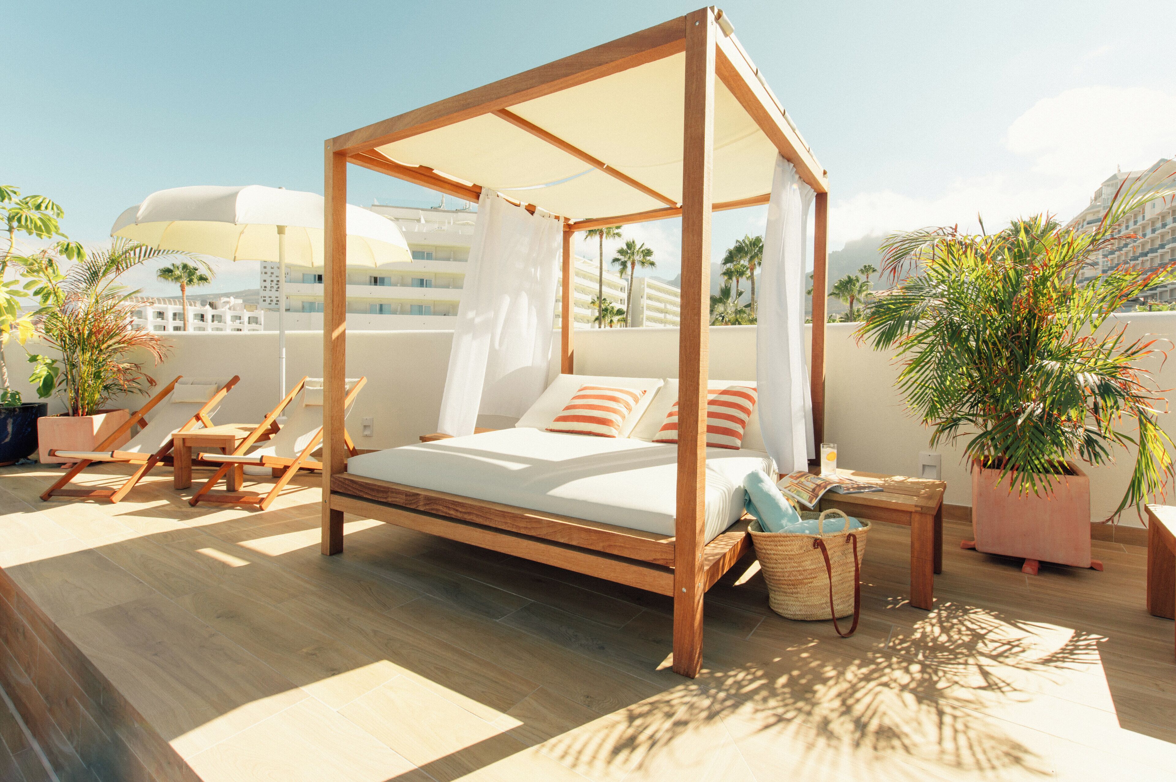 A wooden four-poster bed with white bedding and orange stripes, surrounded by lounge chairs and potted plants on a sunlit terrace with a view of palm trees and a building in the background.