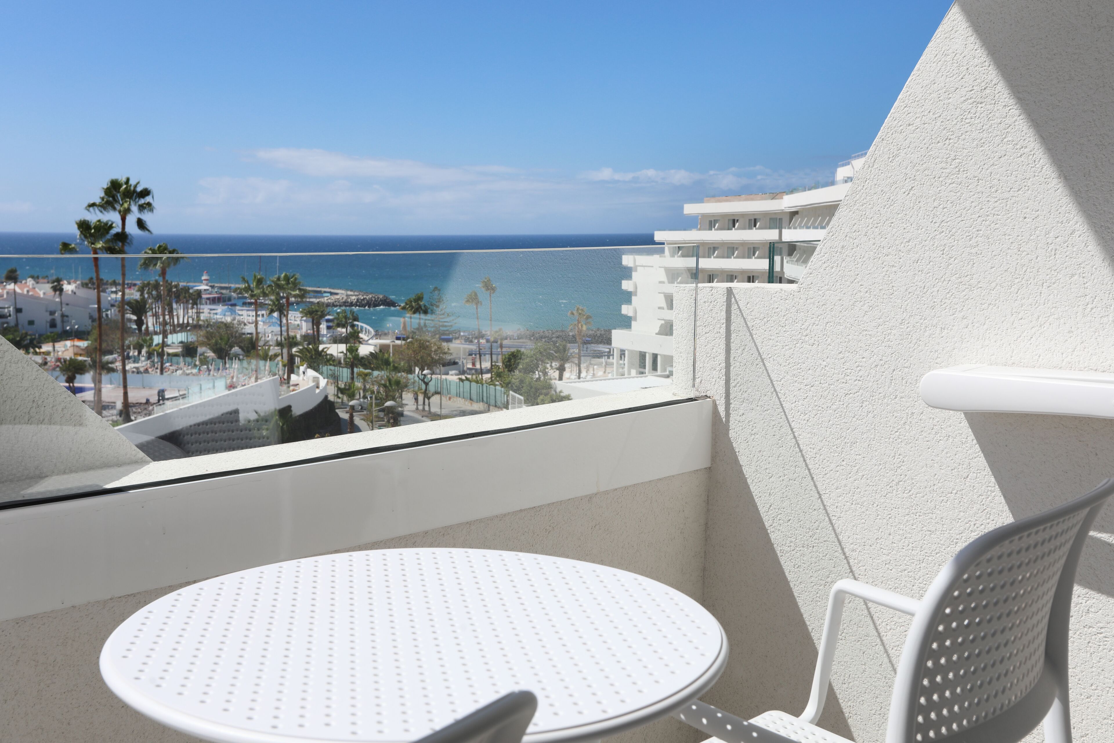 Un balcón con una mesa y una silla blancos, con vistas a una playa y una zona turística.