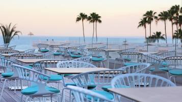 Un café frente al mar con sillas y mesas de metal blanco, dispuestas sobre una terraza de madera, con vista al océano.
