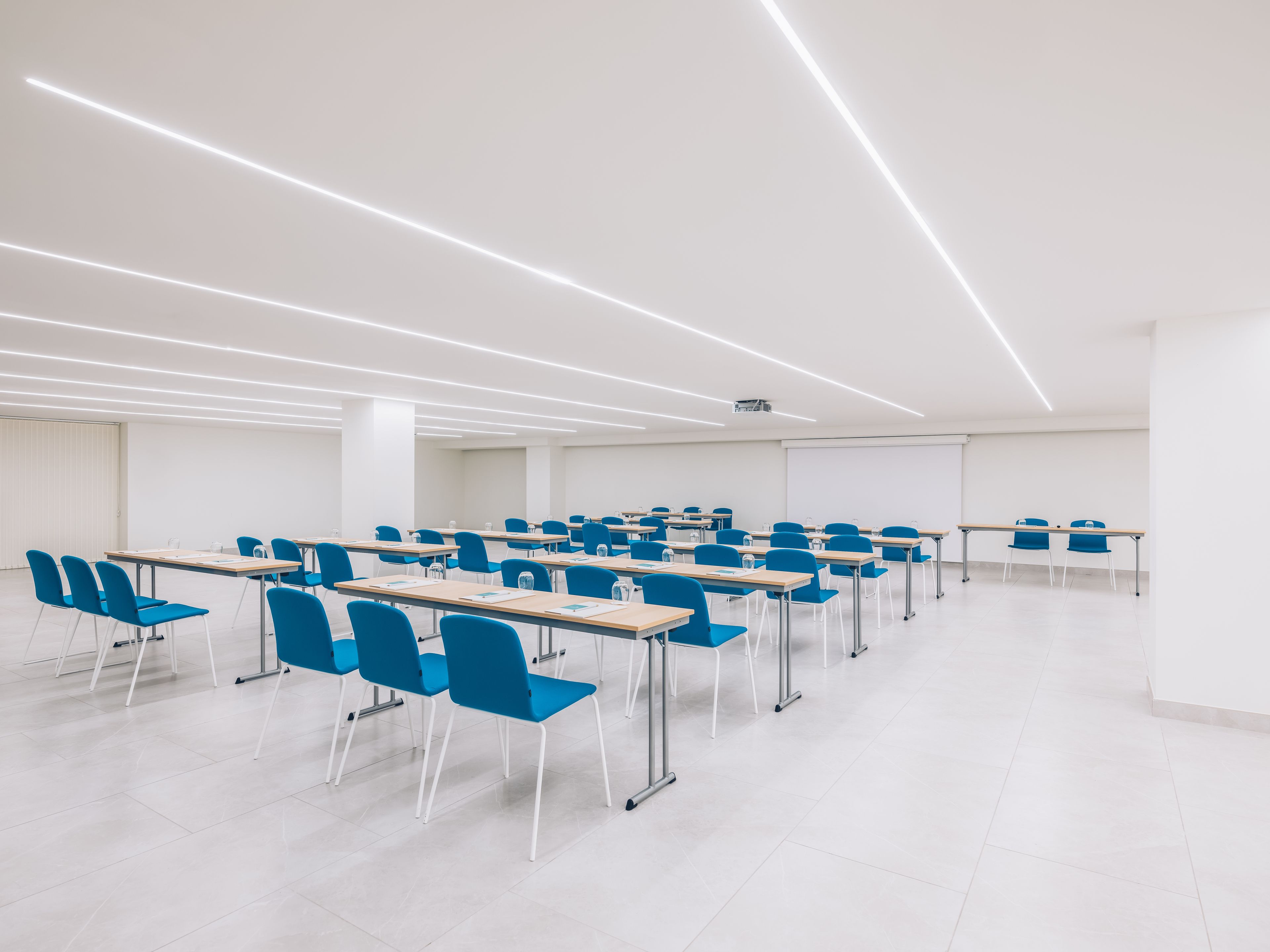 Ein moderner Konferenzraum mit Reihen blauer Stühle und Tische, weißen Wänden und einem Projektor an der Decke.