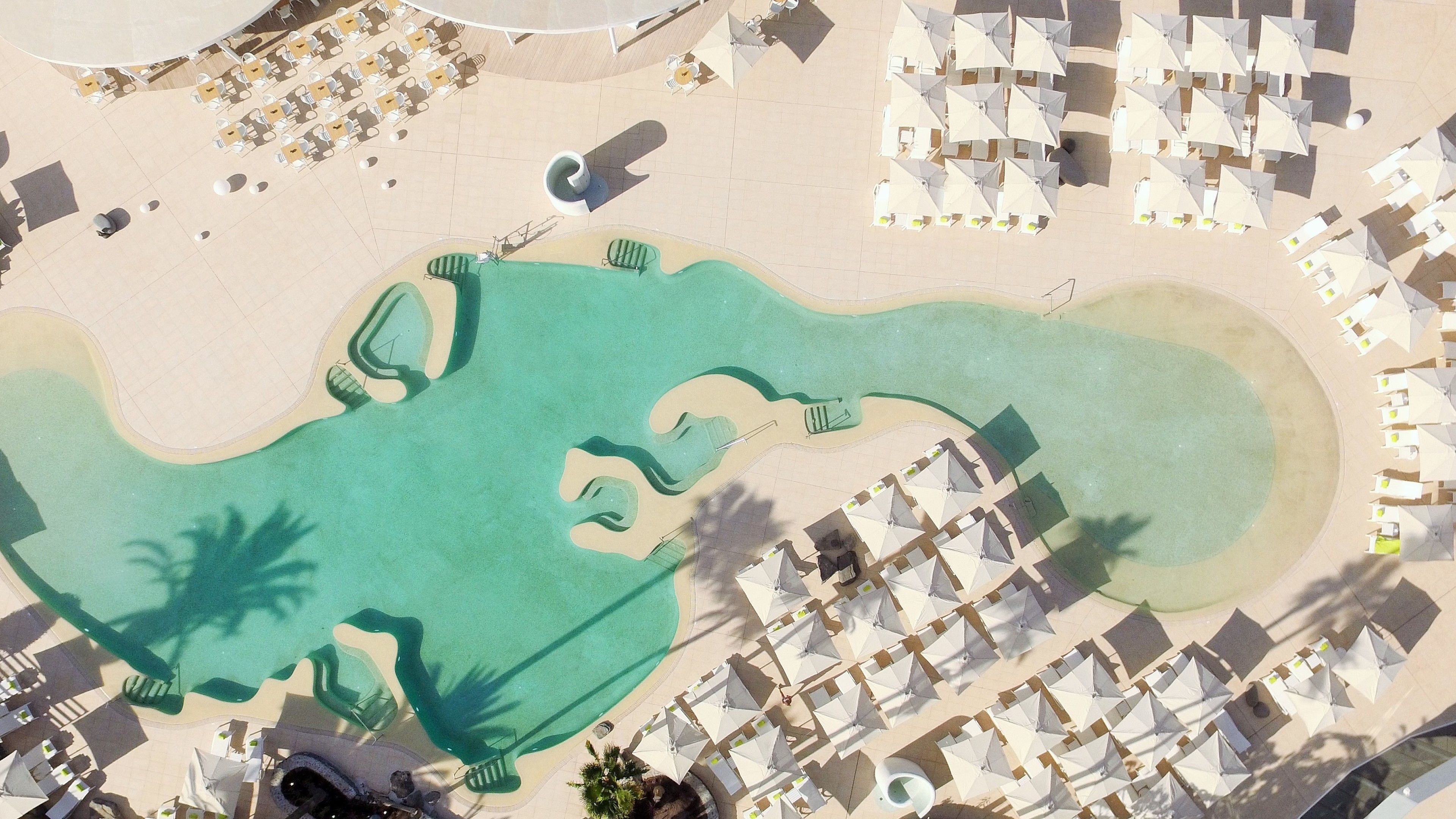 Vista aerea di un resort con una grande piscina, diverse aree relax con ombrelloni e una spiaggia sabbiosa.