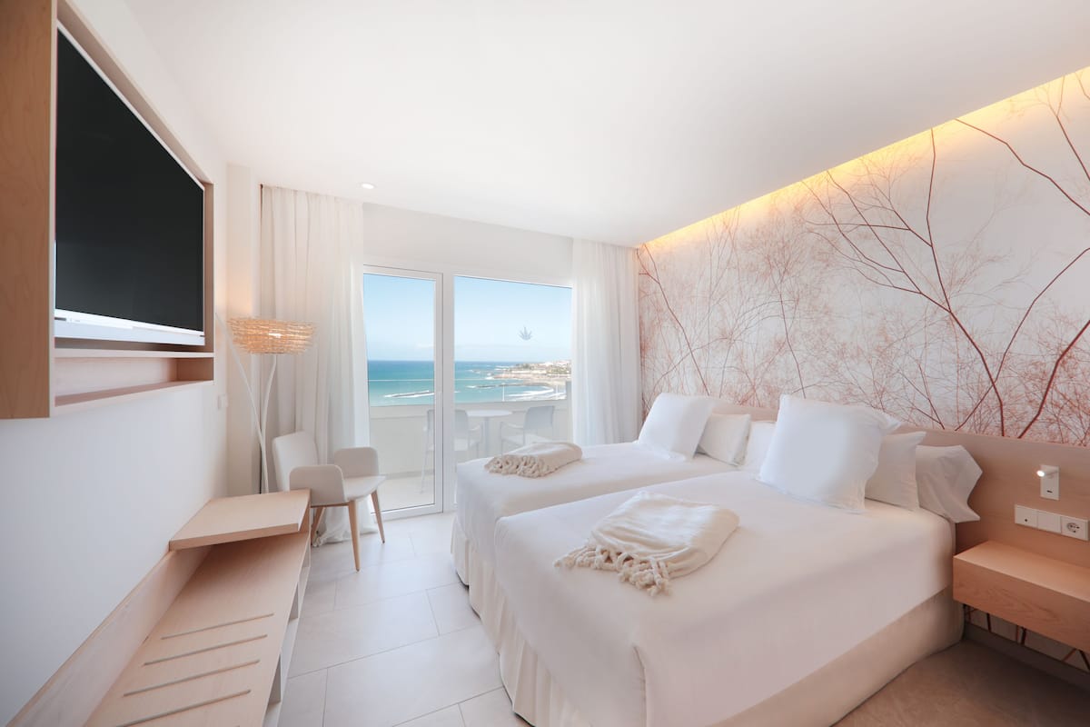 A modern hotel room with two beds, a desk, a chair, and a balcony with a view of the sea.