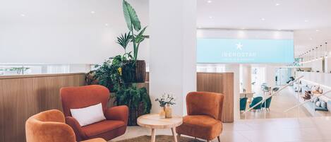 A modern hotel lobby with a reception desk, plush orange seating, a large potted plant, and a visible hotel sign.
