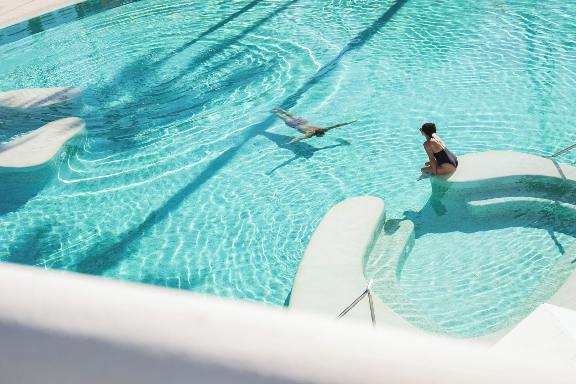 A clear blue pool with a person swimming and another sitting on the edge.