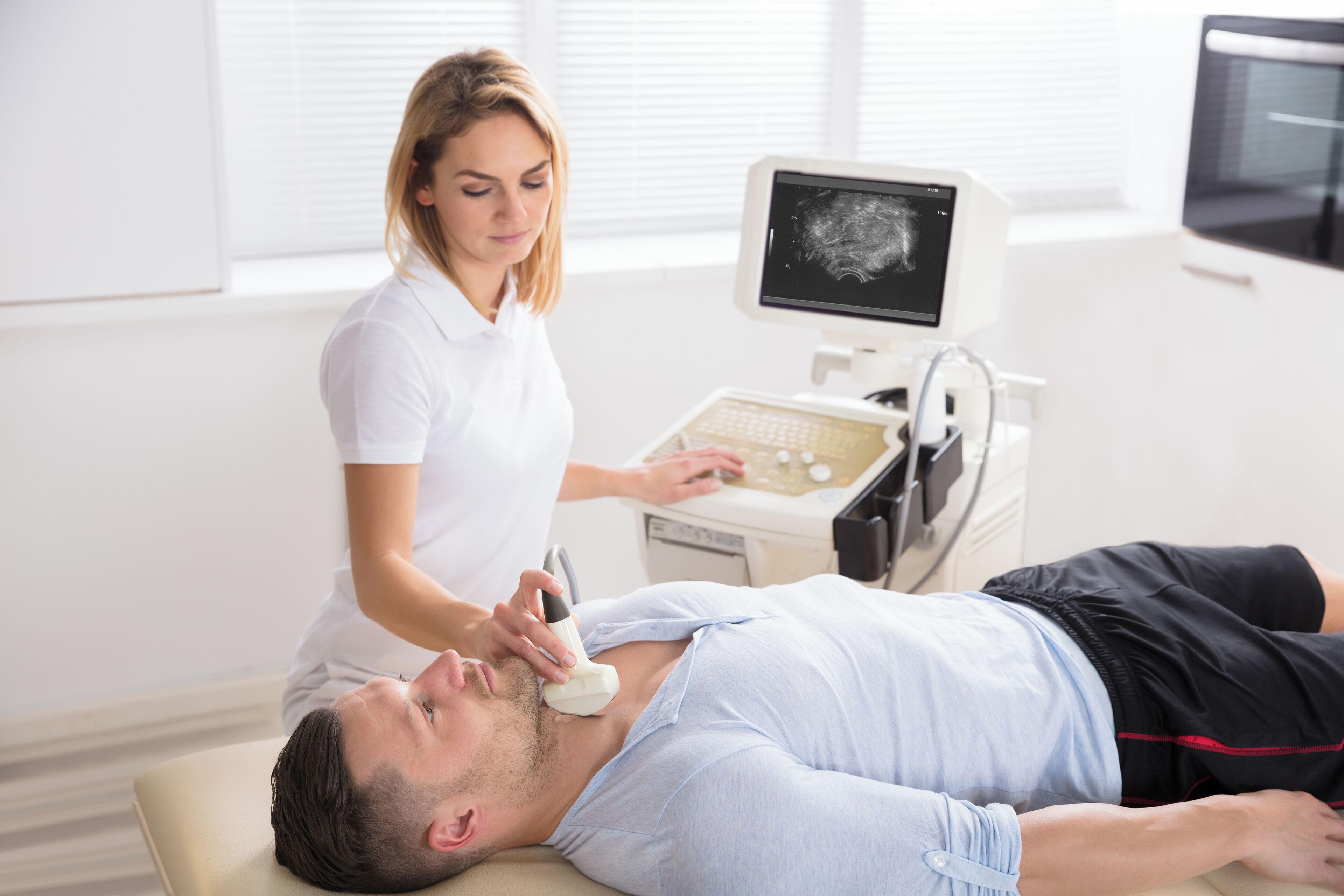 A medical examination room with a patient lying on a bed, a medical professional using a device on the patient, and an ultrasound machine in the background.