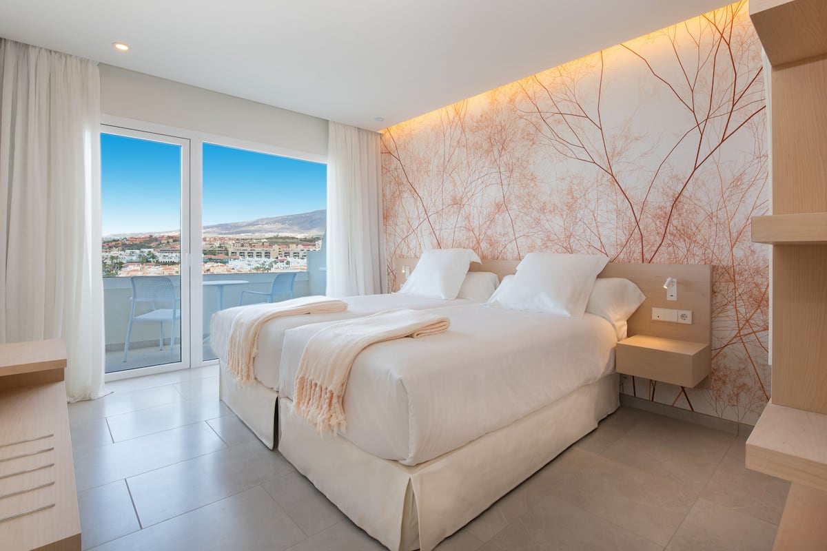 A modern hotel room with a large bed, a bedside table, and a view of the city through the balcony doors.