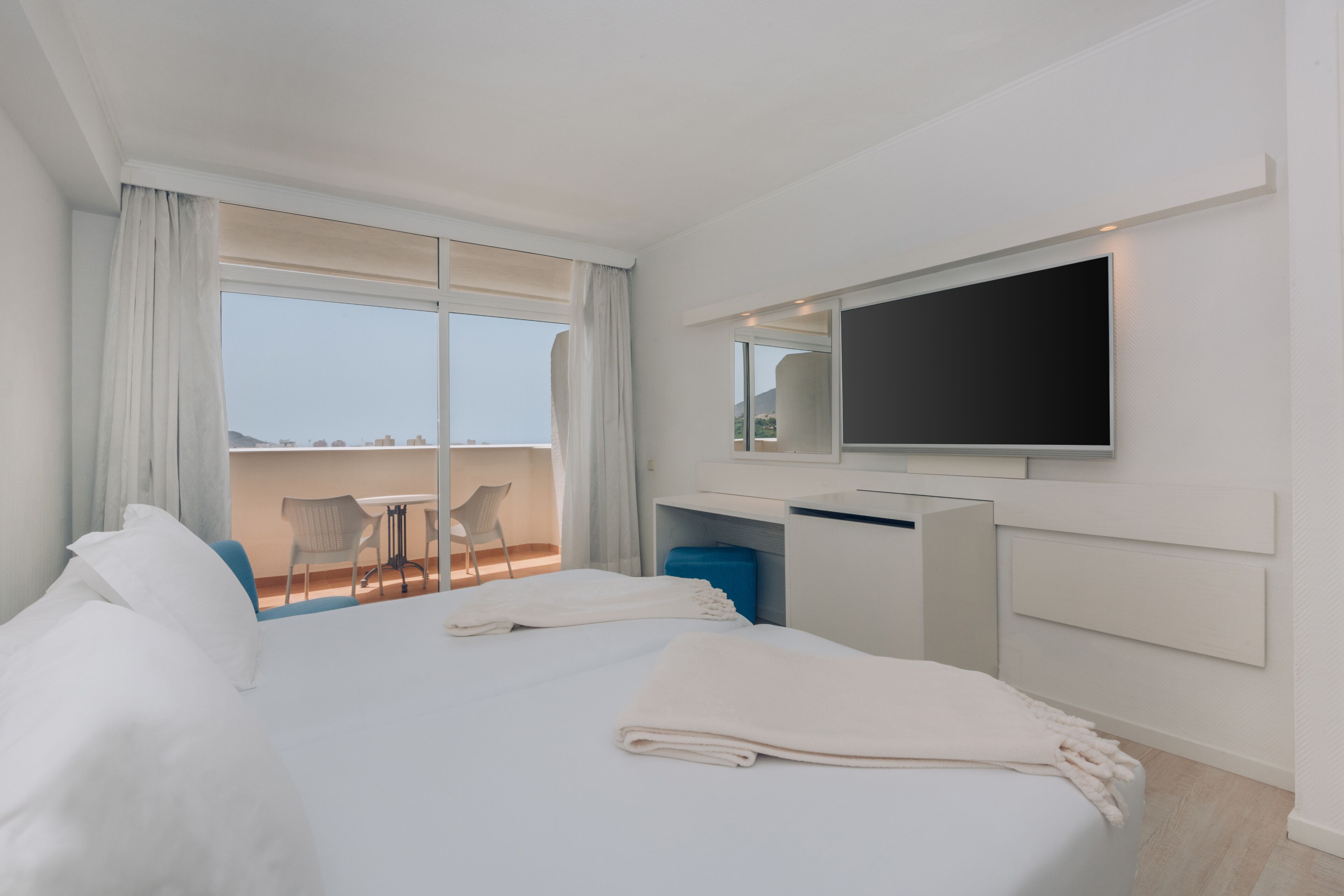 A modern hotel room with a large bed, a TV mounted on the wall, a desk, and a balcony with a view.