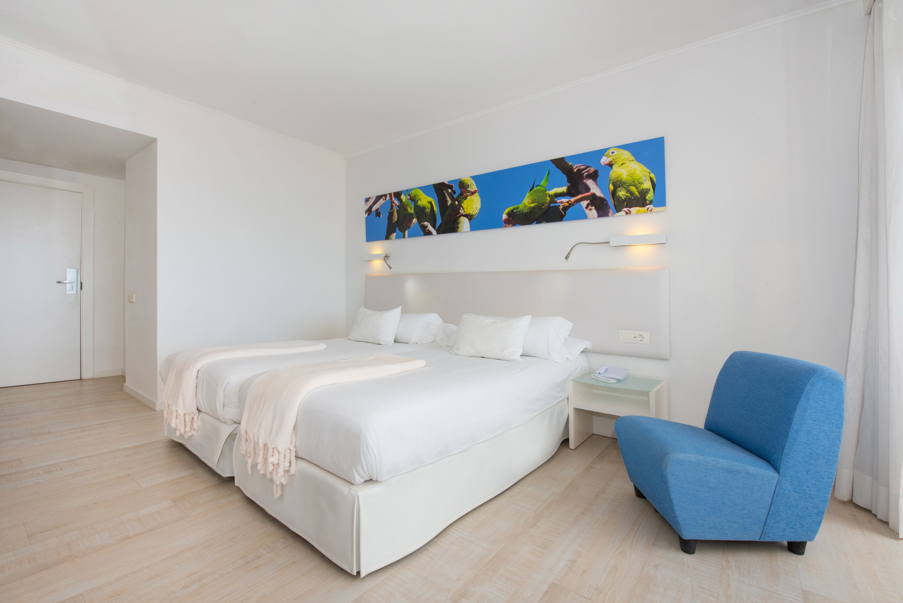 Een moderne hotelkamer met een groot bed, een blauwe fauteuil en een schilderij van papegaaien aan de muur.