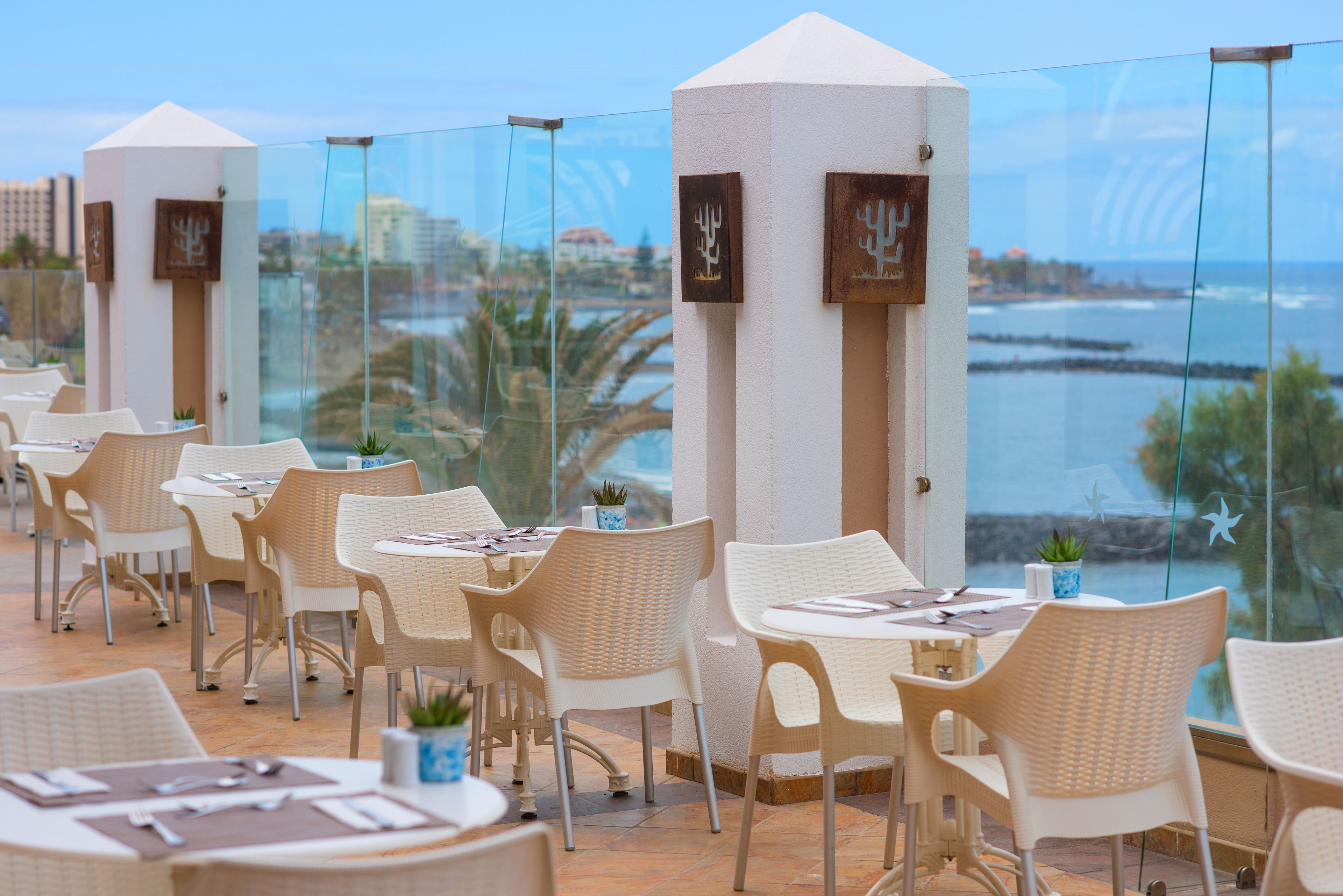 Una terraza con sillas y mesas de mimbre, con vistas a la costa, palmeras y un cielo despejado.