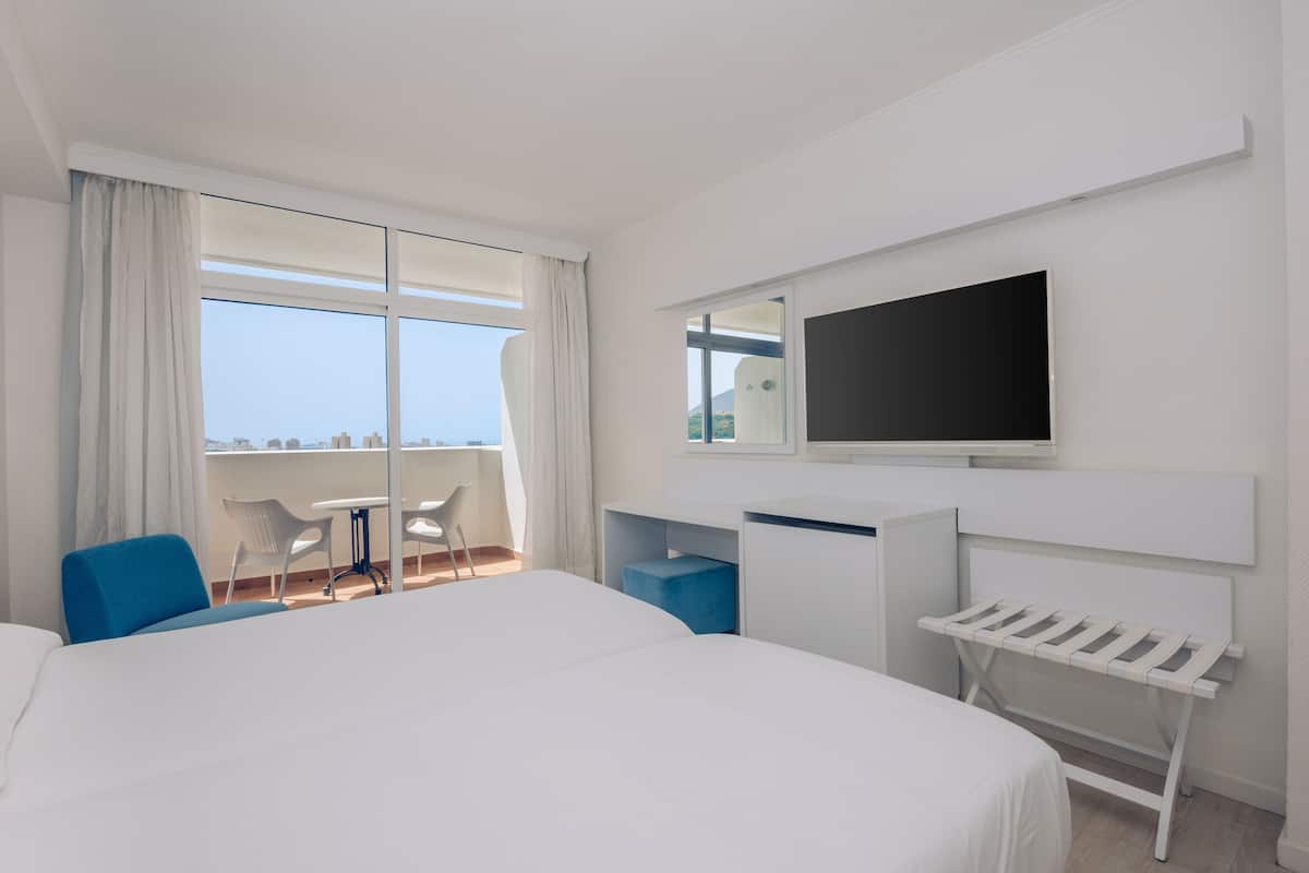 Quarto de hotel moderno com uma cama grande, TV fixada na parede, uma mesinha com cadeiras e varanda com vista.