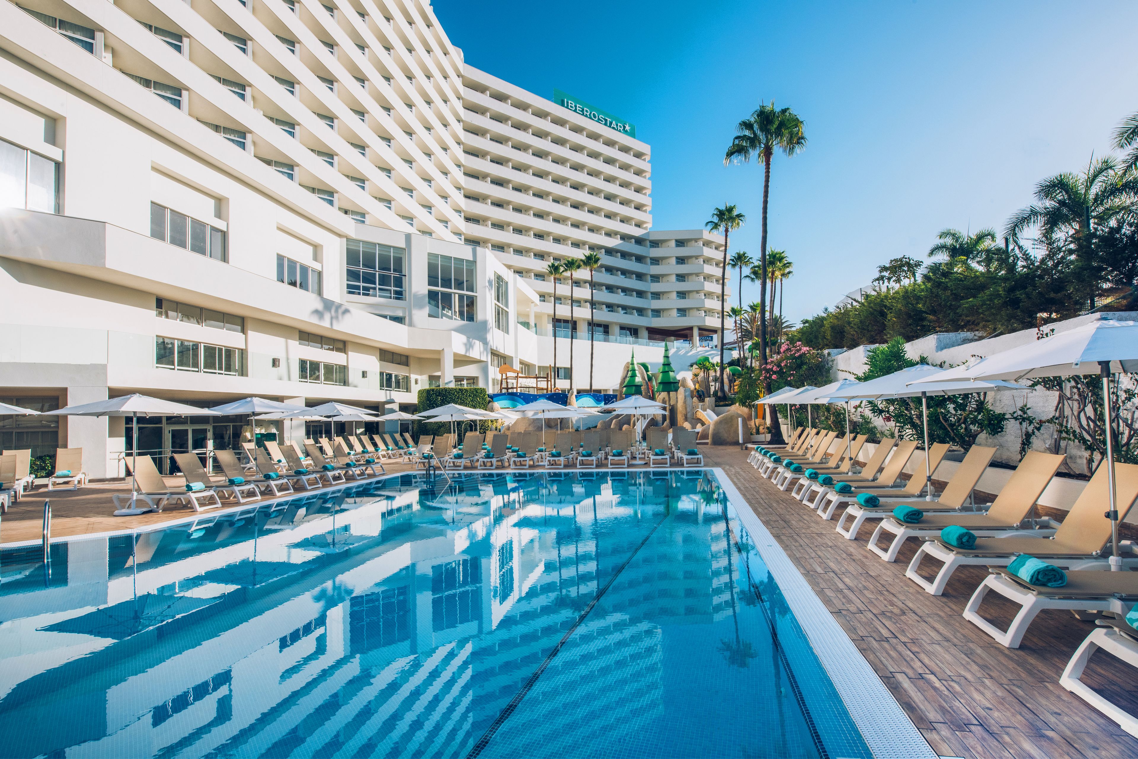 Zona de piscina con tumbonas y sombrillas, un edificio grande con balcones y palmeras.