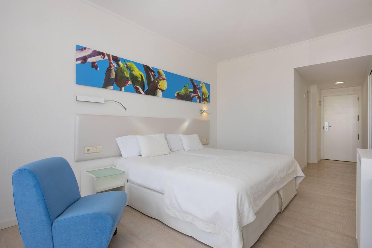 Quarto de hotel com uma cama grande, uma poltrona azul, uma mesa pequena e uma obra de arte na parede.