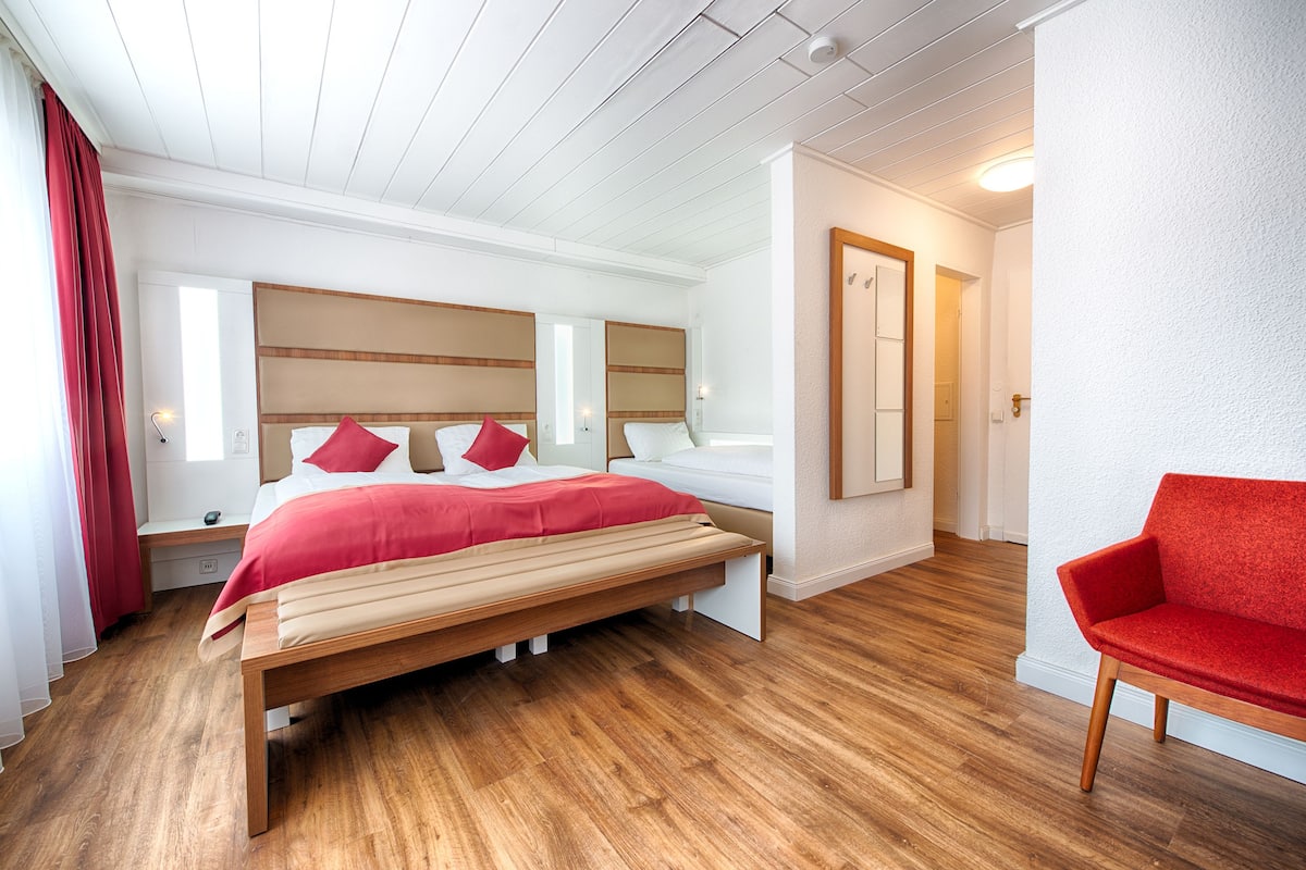 A bedroom with a large bed, a wooden bench, a red chair, and a mirror on the wall.