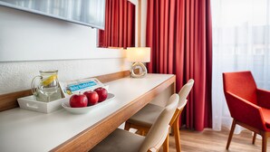 Habitación de hotel con un escritorio de madera, una silla roja, una lámpara blanca, una jarrón de vidrio con un limón y un plato de manzanas.
