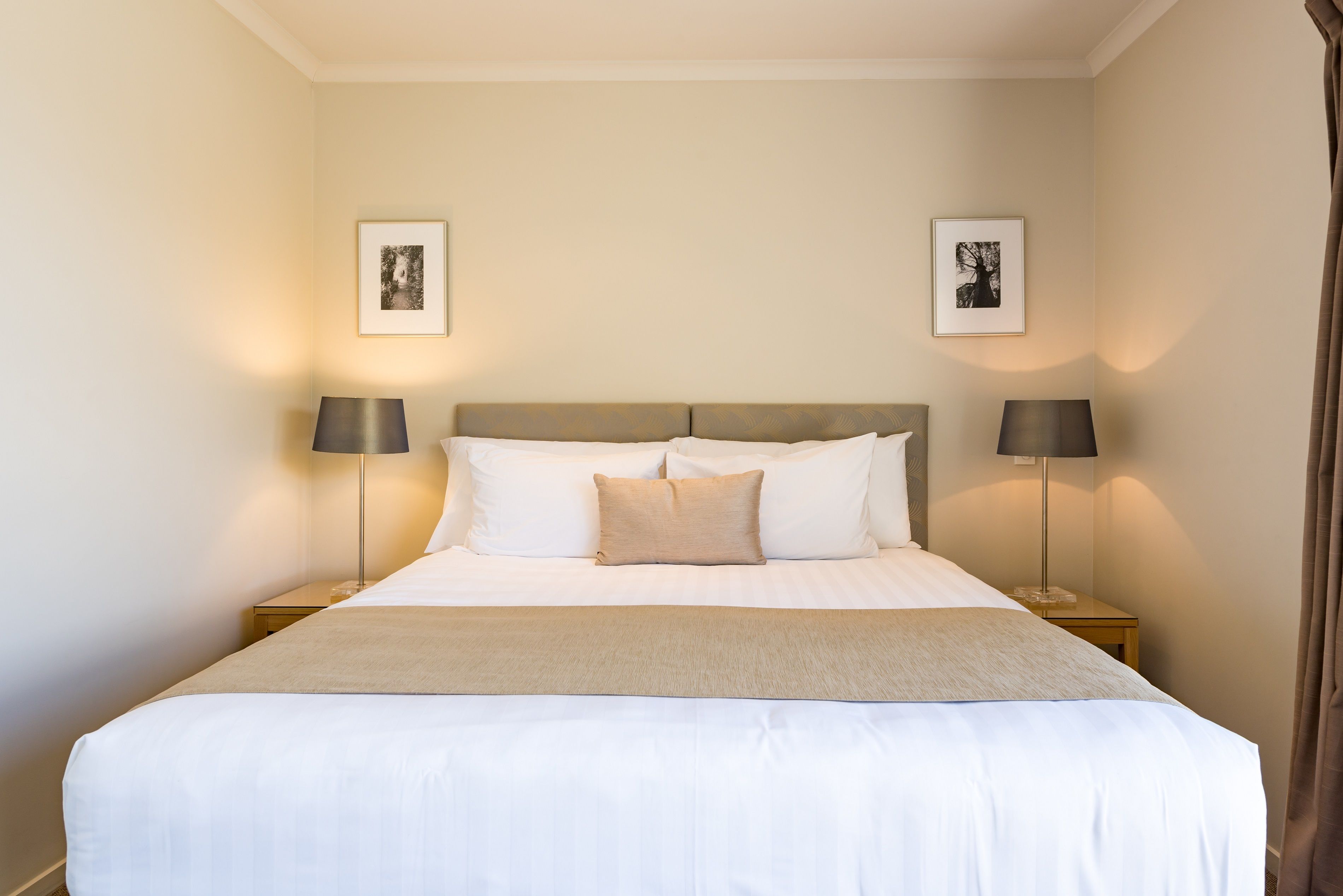 Una cama bien tendida con sábanas blancas y una manta color beige, con dos lámparas de noche a cada lado y cuadros en las paredes.