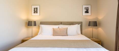 Una cama bien tendida con sábanas blancas y una manta color beige, con dos lámparas de noche a cada lado y cuadros en las paredes.