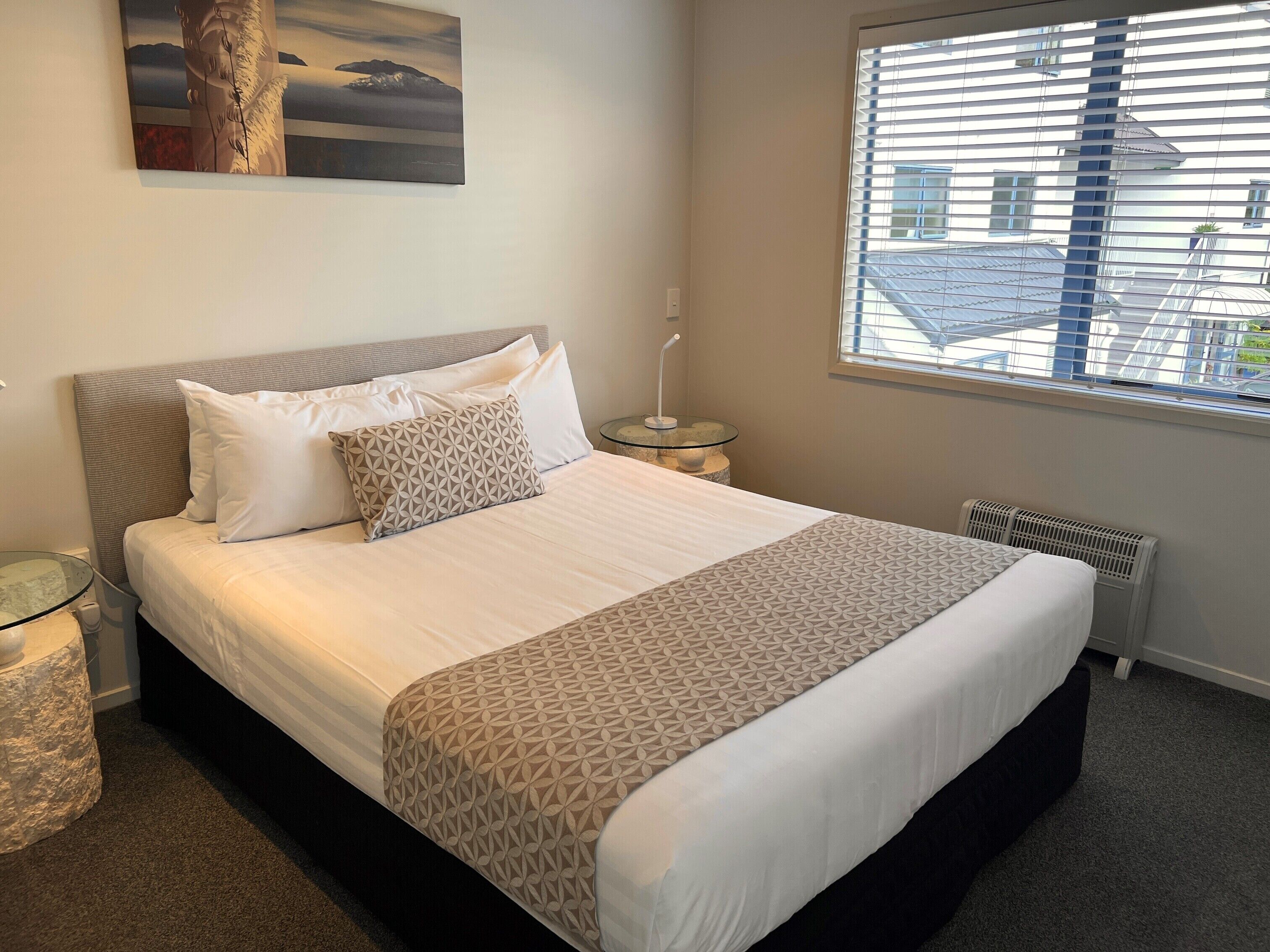 A neatly made bed with a patterned bedspread and pillows, a glass side table with a lamp, a painting on the wall, and a window with blinds.