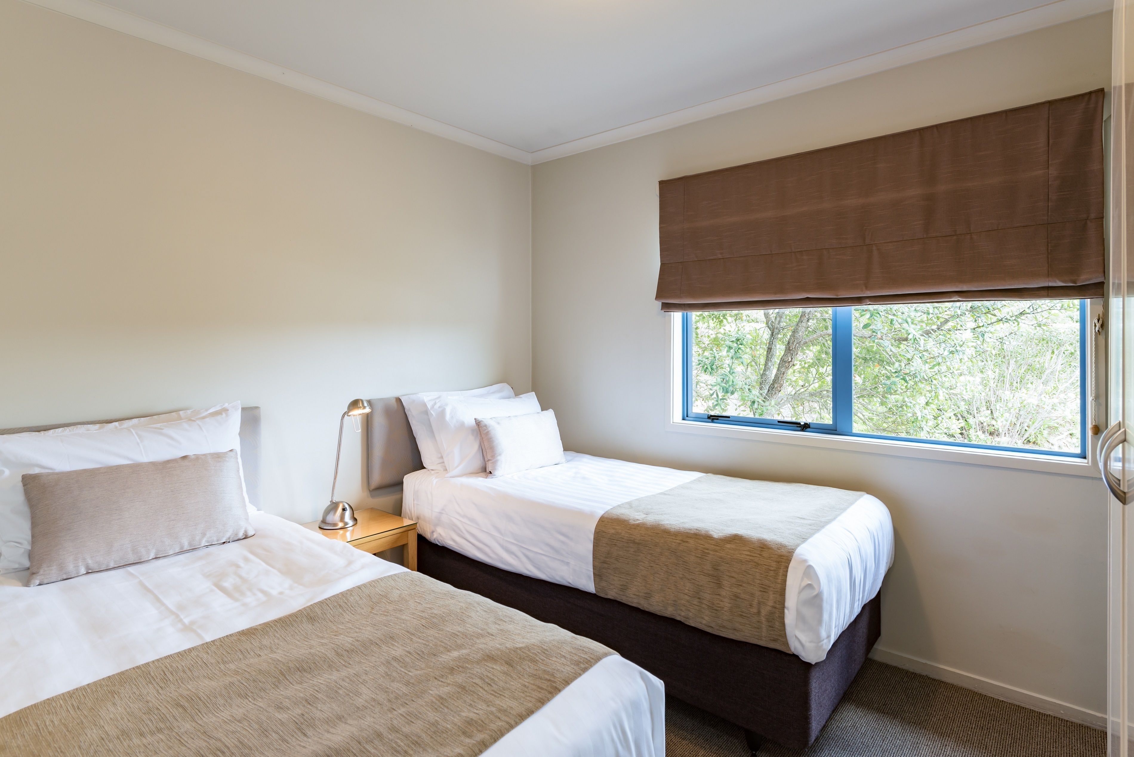 A bedroom with two beds, a window with a view of greenery, and a bedside table with a lamp.
