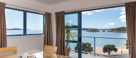 A room with a dining table, chairs, and a large sliding glass door leading to a balcony with a view of the sea and islands.