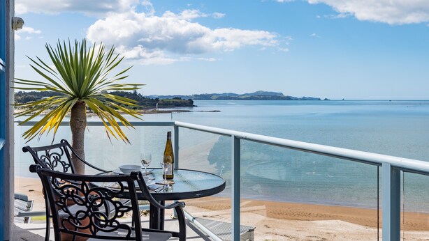 Un balcón con mesa, sillas y una planta de palma con vistas a una playa y al agua.