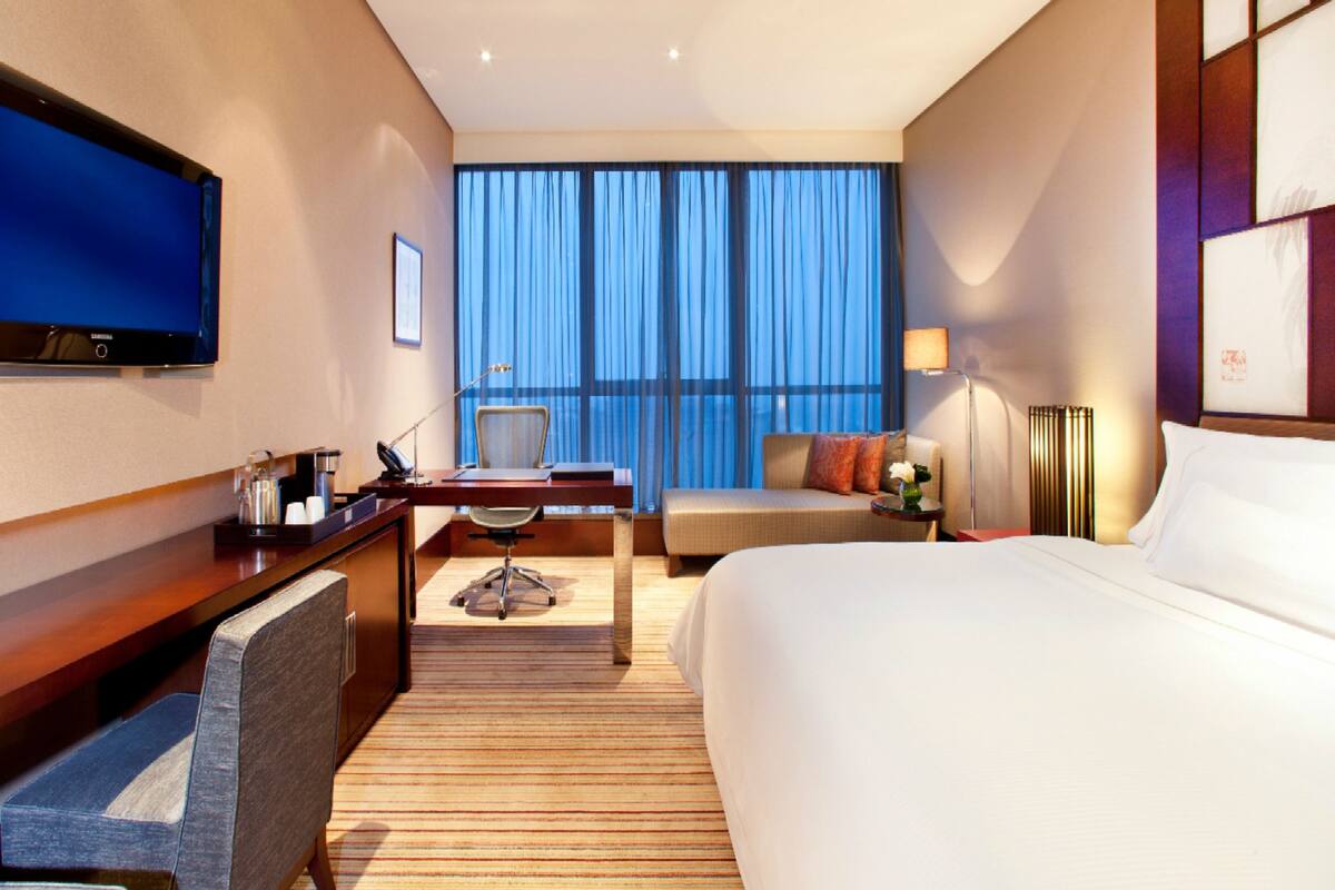 A hotel room with a large bed, a desk with a chair, a television, and a view of the city through large windows.