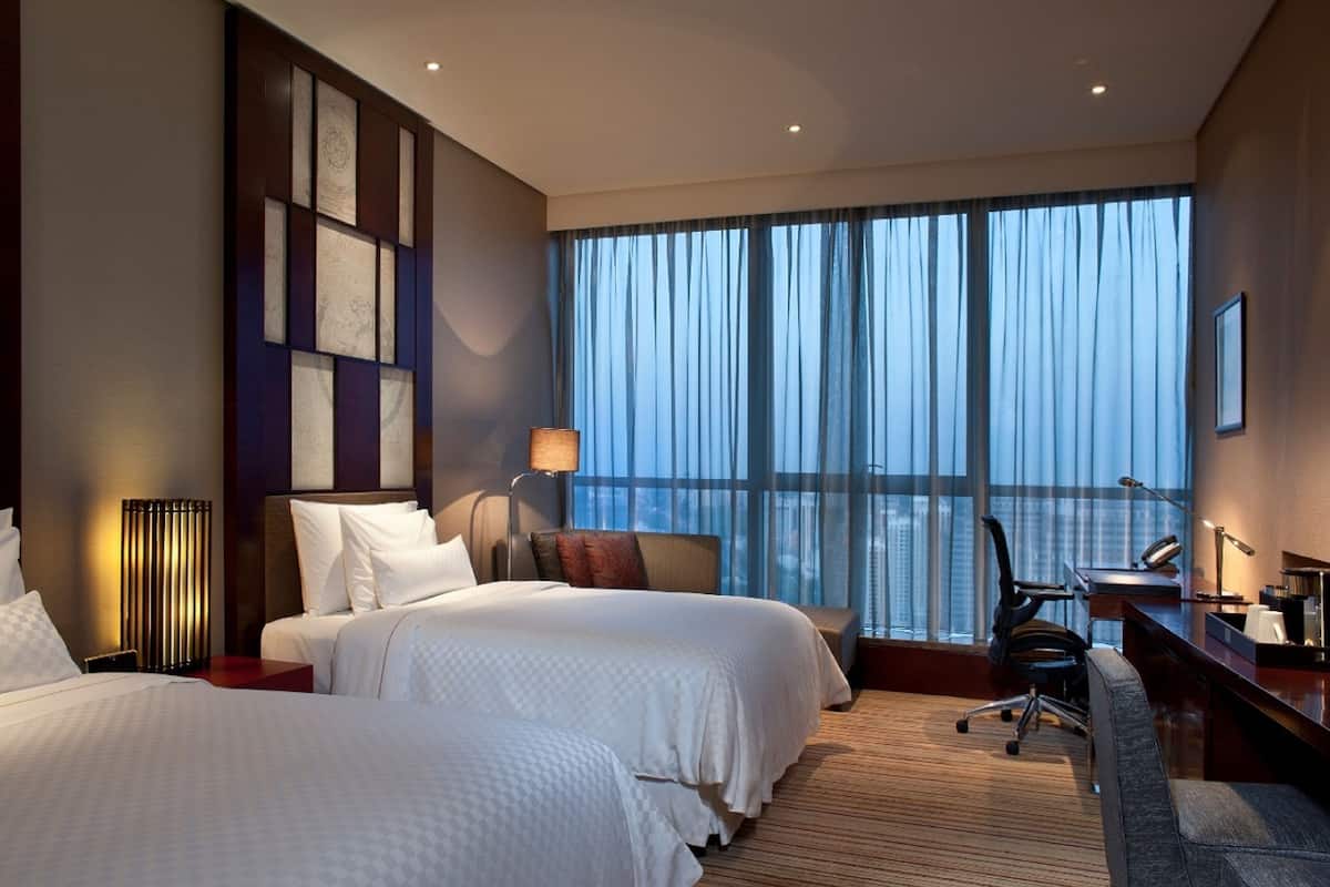 A hotel room with two beds, a desk, a chair, and a large window with curtains.