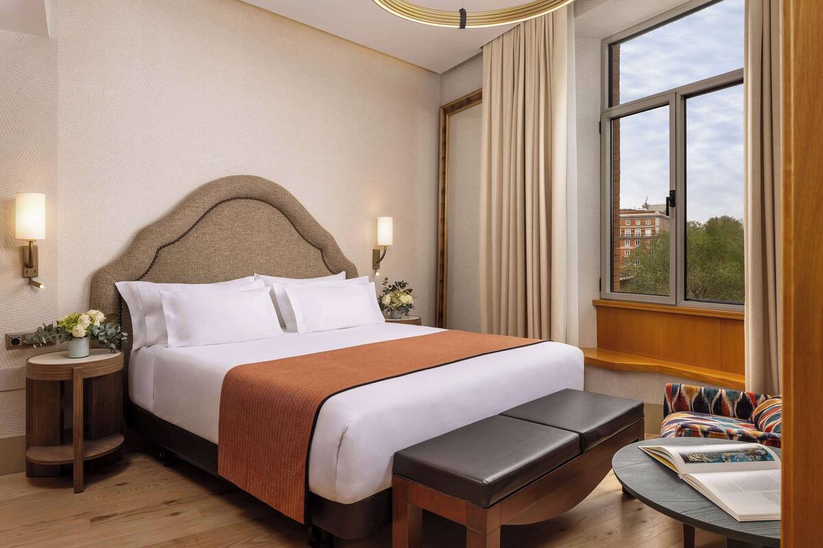 A hotel room with a large bed, a bench, a small table with a book, a nightstand with a lamp, and a window with a view of buildings.