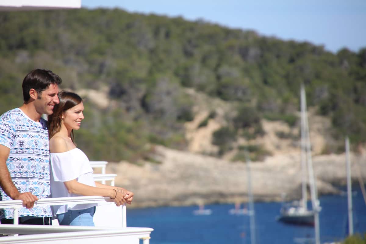 Un couple se tient sur un balcon donnant sur une zone côtière, avec des bateaux et de la végétation en contrebas.