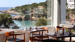 A dining area with tables and chairs, a view of a coastal landscape, and a window overlooking the sea. - Sandos El Greco Hotel - Adults Only (Sant Joan de Labritja)