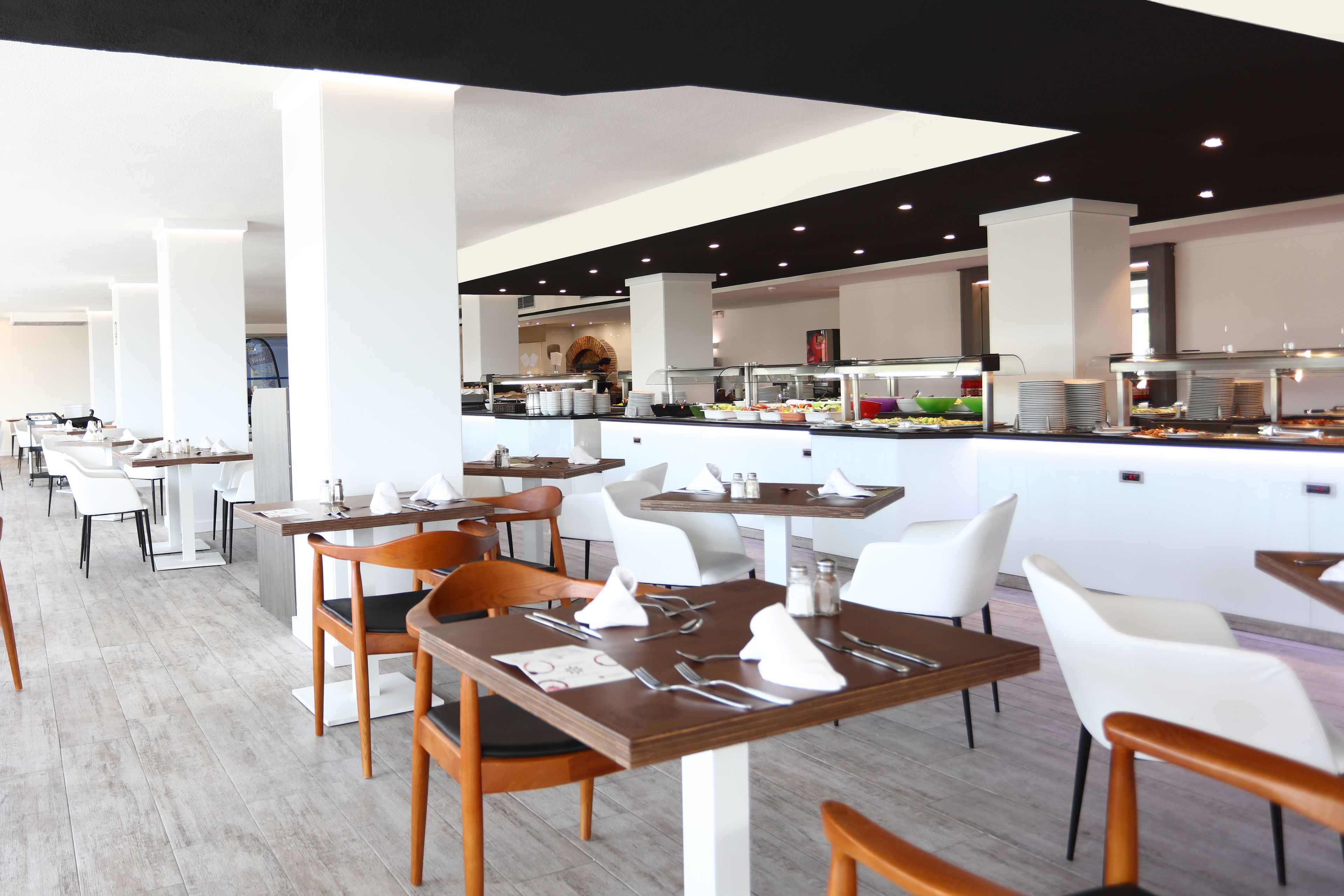 A modern restaurant with white and wooden tables, chairs, and a buffet area in the background.