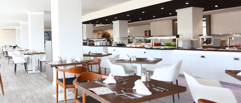 A modern restaurant with white and wooden tables, chairs, and a buffet area in the background.
