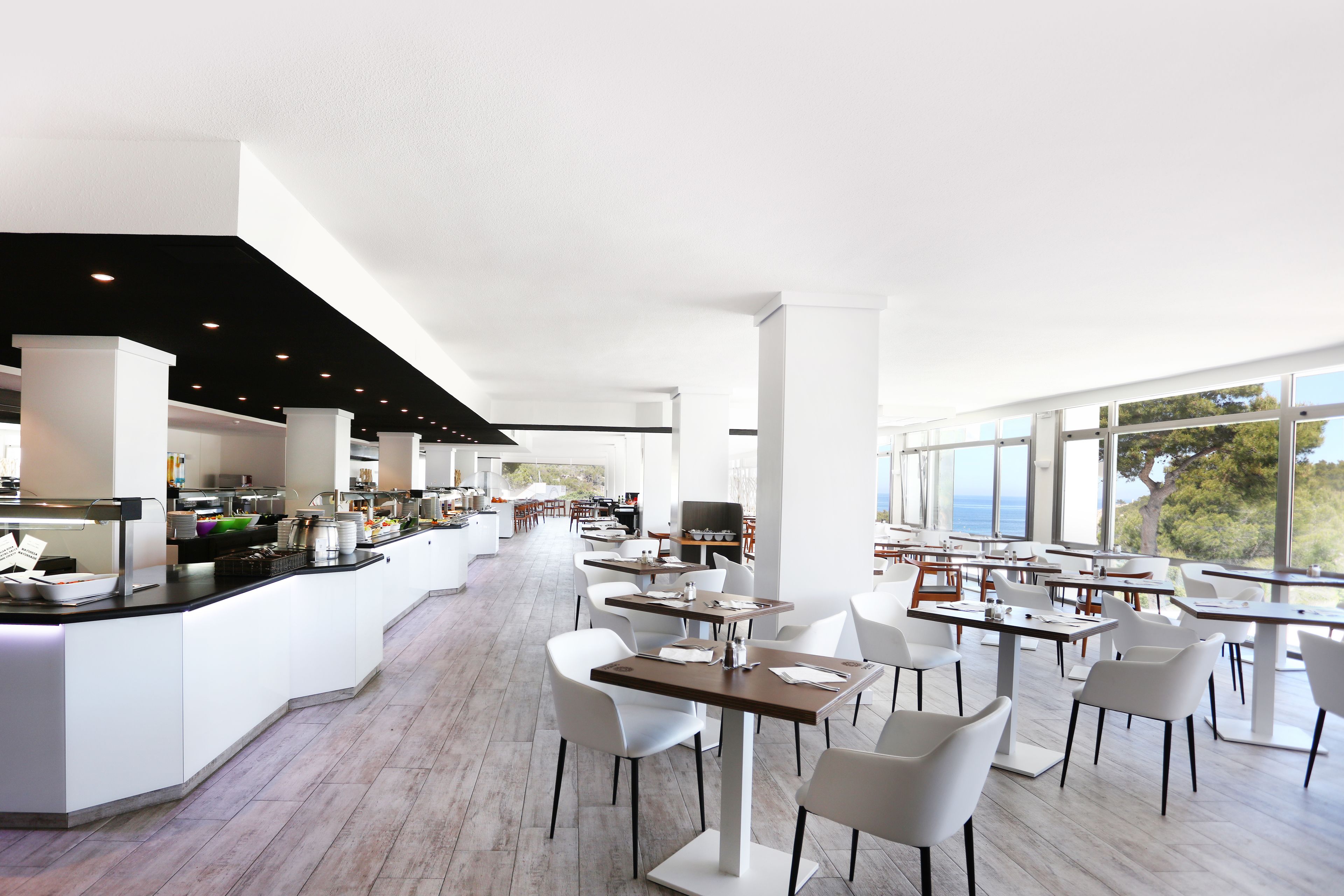 Un restaurante moderno con mobiliario blanco, una barra central y amplios ventanales con vistas al mar.