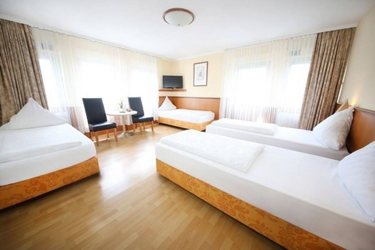 Een hotelkamer met drie enkele bedden, een bureau, een stoel, een televisie en grote ramen met gordijnen.