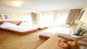 A hotel room with two beds, a wooden floor, a desk with a bowl of fruit, a remote control, and a vase with flowers. - Hotel Europa Offenbach (Offenbach am Main)