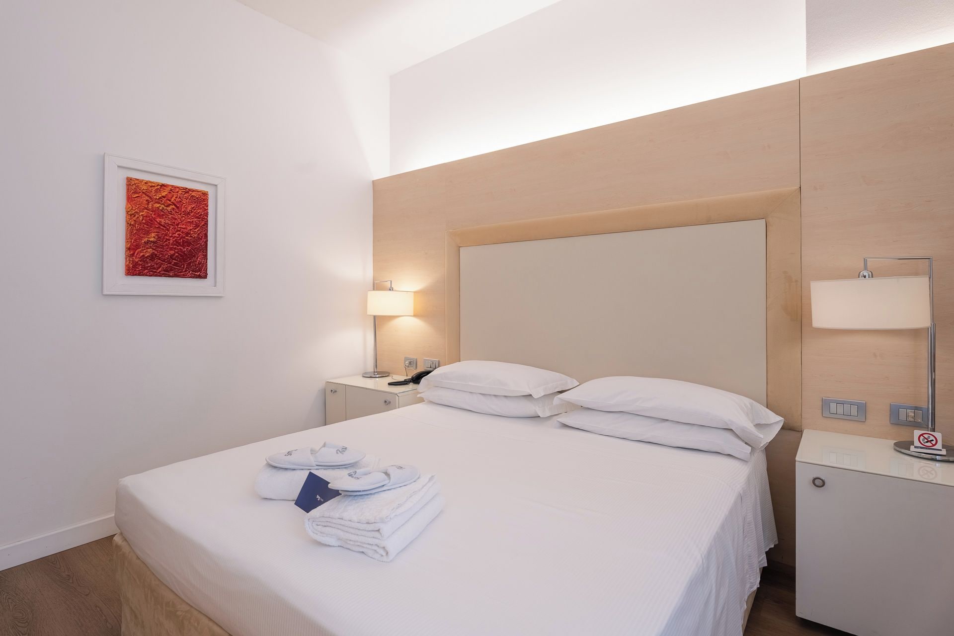 A hotel room with a bed, a nightstand, a lamp, a wall-mounted light, and a framed picture.