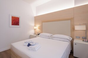 Una habitación de hotel con cama, mesita de noche, lámpara, luz empotrada en la pared y un cuadro enmarcado.