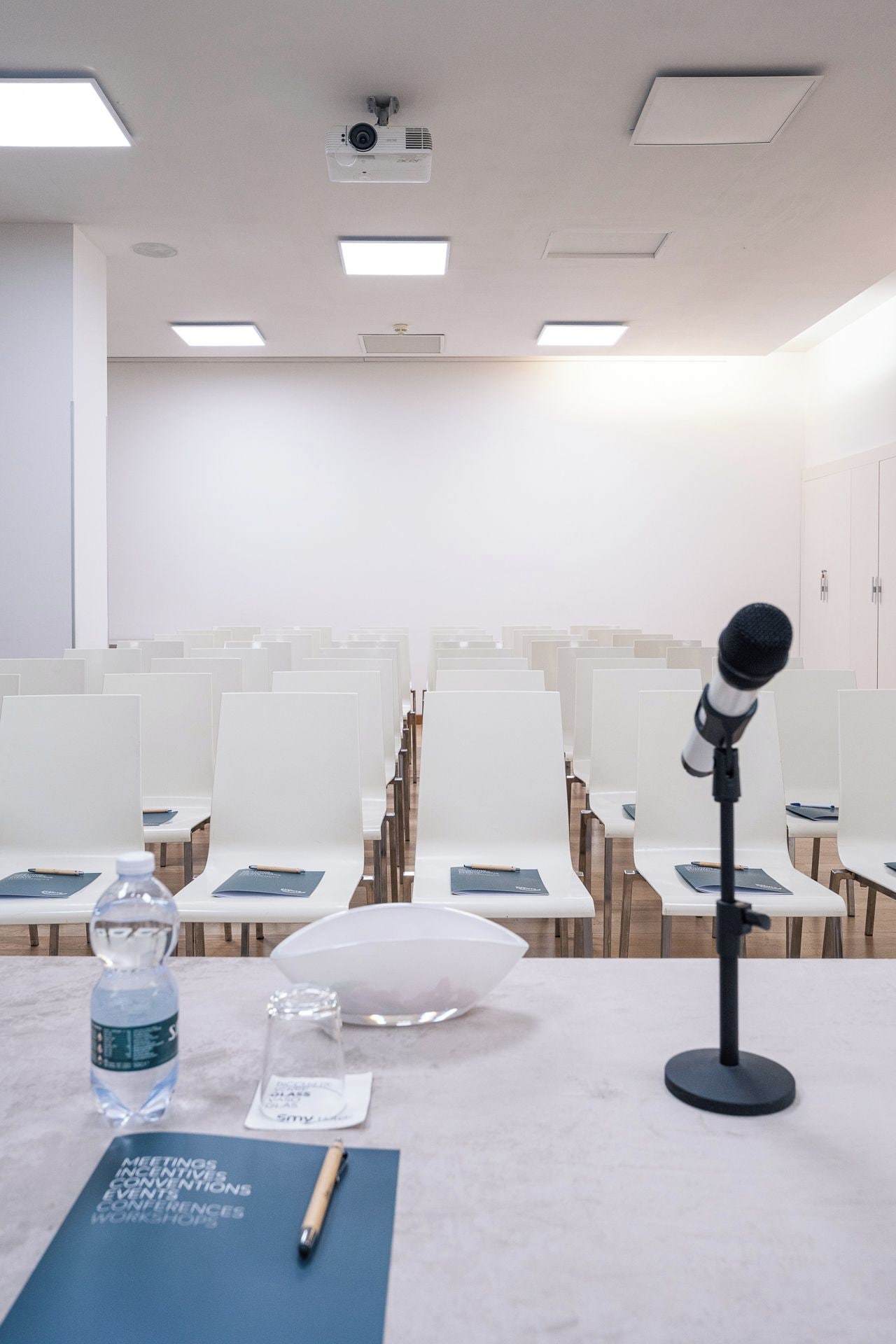 Een vergaderzaal met rijen witte stoelen, een projecter aan het plafond, een microfoon op een statief en een tafel met een waterfles, glas en een notitieboek.