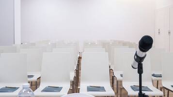 Een vergaderzaal met rijen witte stoelen, een projecter aan het plafond, een microfoon op een statief en een tafel met een waterfles, glas en een notitieboek.
