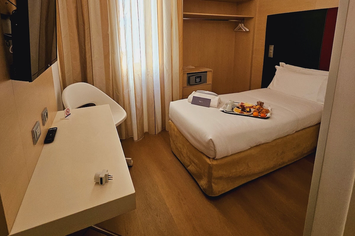 A hotel room with a bed, a desk, a chair, a TV, and a window with curtains.