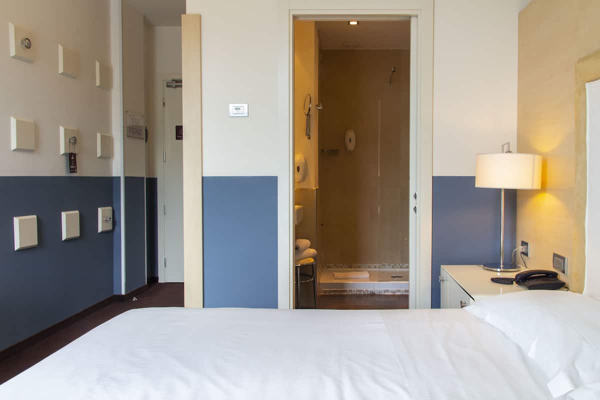 A hotel room with a bed, a bedside table with a lamp, a shower area, and a door leading to another room.